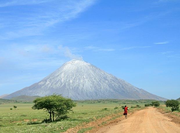 Flocks of thousands of bright pink lesser flamingos wading across the glass-like crystalline surface of Lake Natron with Ol Doinyo Lengai volcano towering behind
